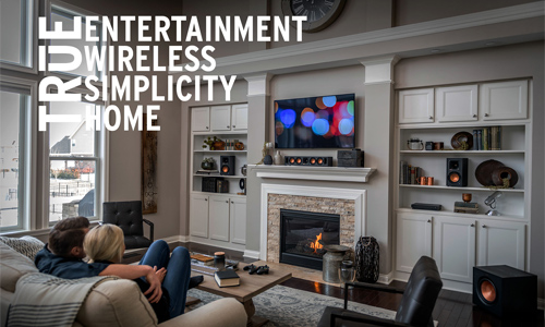Living room scene with Klipsch products setting on built-in shelves and the subwoofer in the corner on the floor and True Entertainment, Wirless, Simplicity, and Home text overlayed on image