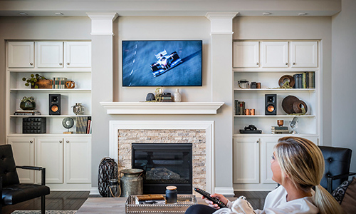 Woman sitting on sofa holding remote and watching TV with Klipsch products placed on built-in shelves in room